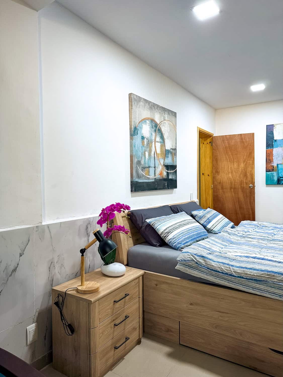 Pohled na moderní pokoj s postelí, nočním stolkem a obrazem na stěně.