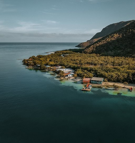 Pohled na klidné moře a pláž u Planet Patanemo s malými domky a zelení.